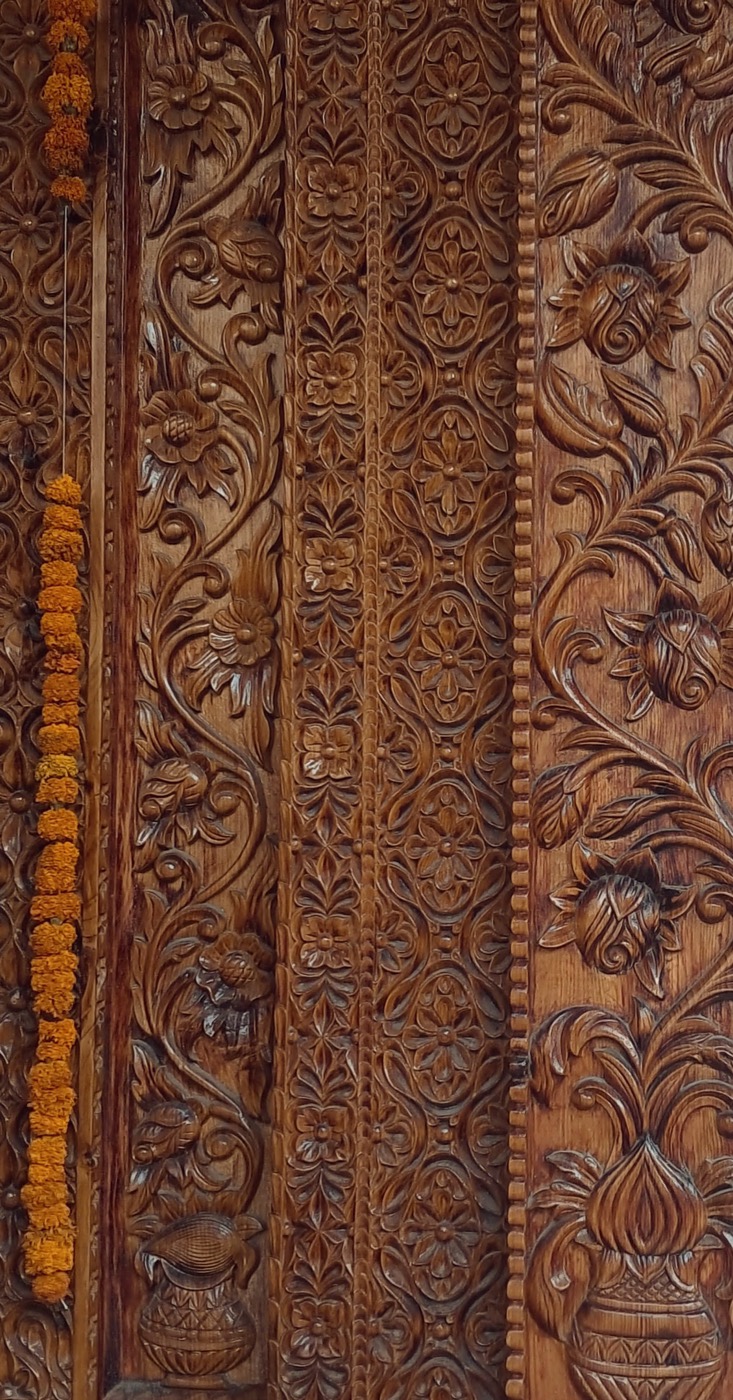 Dense floral carving across a timber door panel, with a marigold garland hanging beside it