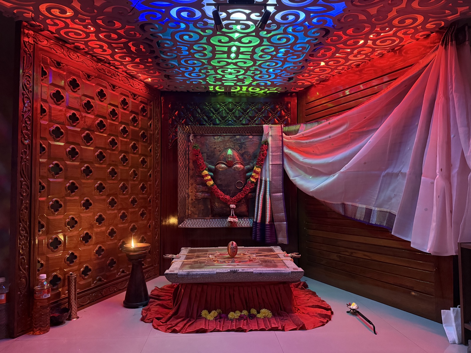 A custom-built pooja room with a carved and pierced timber ceiling and wall panels, a stone altar, and a deity idol dressed with garlands