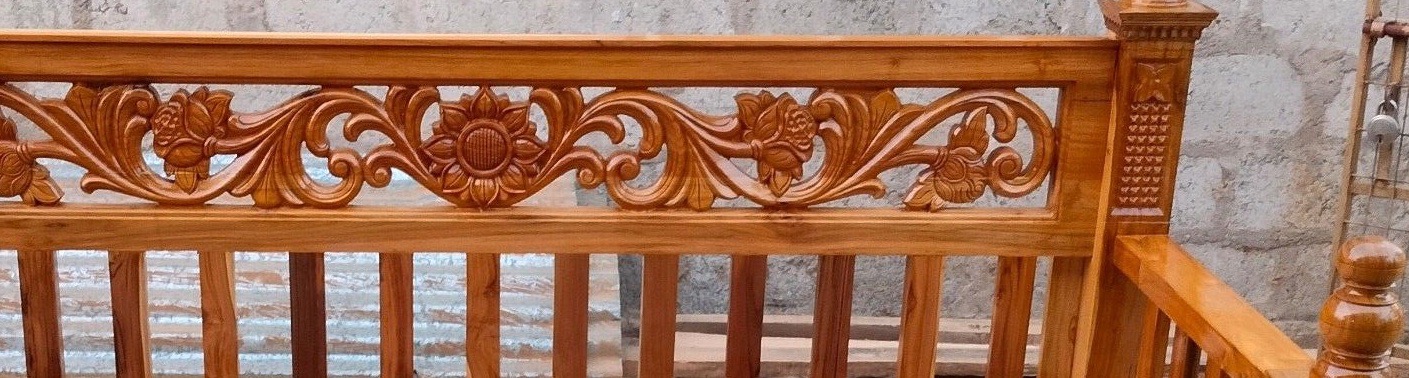 A hand-carved teak headboard rail with floral scrollwork and a central rosette, standing in the workshop